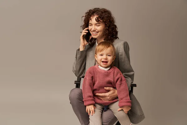 Збалансований спосіб життя, телефонний дзвінок, щаслива робоча мати розмовляє на смартфоні і сидить на стільці з дитиною на сірому фоні, материнство, багатозадачність, якісний час, концепція балансу життя — стокове фото