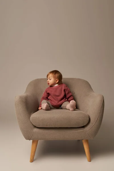 Cute baby girl, toddler in casual attire sitting on comfortable armchair on grey background in studio, emotion, confused, innocence, little child, toddler fashion, stylish outfit, sweater — Stock Photo