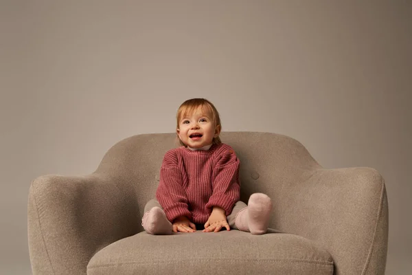 Cute baby girl, toddler in casual attire sitting on comfortable armchair on grey background in studio, emotion, happiness, joy, innocence, little child, toddler fashion, stylish outfit, sweater — Stock Photo