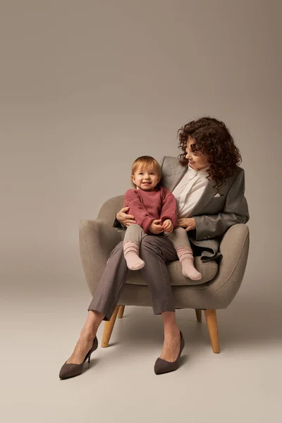 Сучасний працездатний батько, балансуюча робота і концепція життя, кучерява жінка в костюмі сидить у кріслі з дочкою малюка, сірий фон, щаслива мати і дитина, багатозадачність, якісний час — стокове фото
