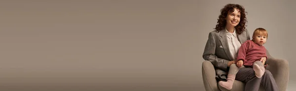 Pai trabalhador moderno, equilíbrio trabalho e conceito de vida, mulher encaracolado em terno sentado em poltrona com a filha da criança, fundo cinza, mãe alegre e criança, multitarefa, tempo de qualidade, banner — Fotografia de Stock
