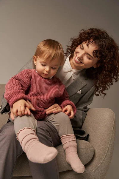 Работающая мать, балансирующая работа и жизненная концепция, кудрявая женщина в костюме, сидящая в кресле с дочкой-малышкой, серый фон, счастливый родитель и ребенок, многозадачность, качественное время — стоковое фото