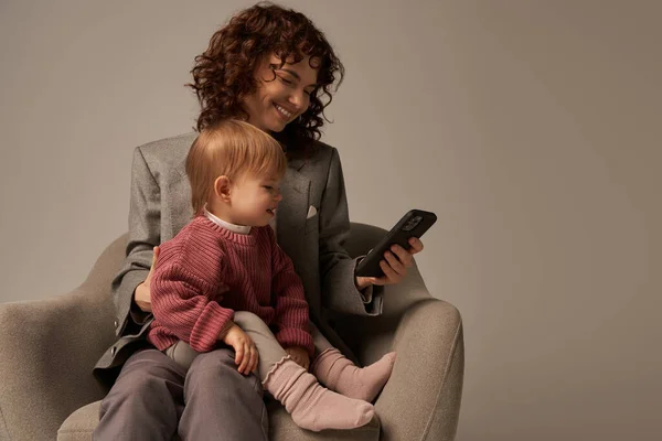 Современный работающий родитель, балансировка работы и концепции жизни, счастливая женщина в костюме сидя с дочкой малыша, используя смартфон, серый фон, счастливая мать и ребенок, многозадачность — стоковое фото