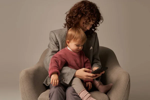 Многозадачность, пользователь смартфона, современная работающая мать, балансирующая работа и жизненная концепция, кудрявая женщина в костюме, сидящая с дочкой на кресле, серый фон, родитель и ребенок — стоковое фото