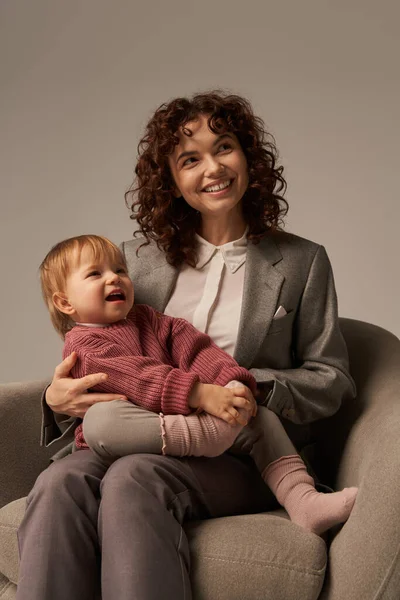 Modern working mother, balancing work and life concept, happy businesswoman in suit sitting with toddler daughter on armchair, engaging with child, grey background, family relationships — Stock Photo