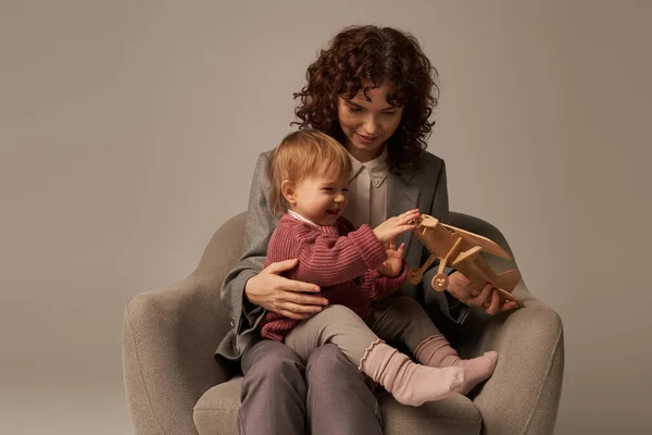 Modern working mother, balancing work and life concept, businesswoman in suit sitting on armchair and playing with toddler daughter, wooden biplane, grey background, engaging with child — Stock Photo