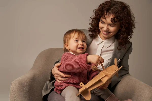 Career and family, balancing work and life concept, businesswoman in suit sitting on armchair and playing with toddler daughter, wooden biplane, grey background, engaging with child — Stock Photo