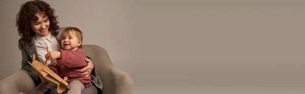 Moderne berufstätige Mutter, Spagat zwischen Arbeit und Lebenskonzept, Geschäftsfrau im Anzug auf Sessel sitzend und mit Kleinkind spielend, hölzerner Doppeldecker, grauer Hintergrund, kindgerecht, Banner — Stockfoto