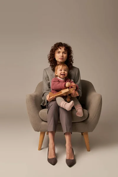 Кар'єра та сім'я, балансування роботи та концепції життя, ділова жінка в костюмі сидить на кріслі з дочкою малюка, грає з дерев'яним біплан, сірий фон, материнство, повна довжина — стокове фото