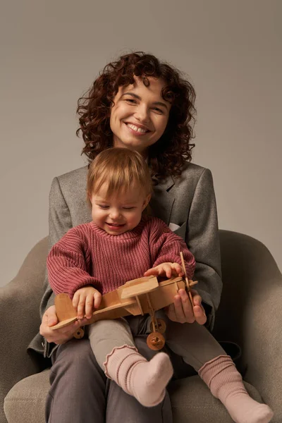 Modern working parent, balancing work and life concept, businesswoman in suit sitting on armchair and playing with toddler daughter, wooden biplane, grey background, engaging with child — Stock Photo