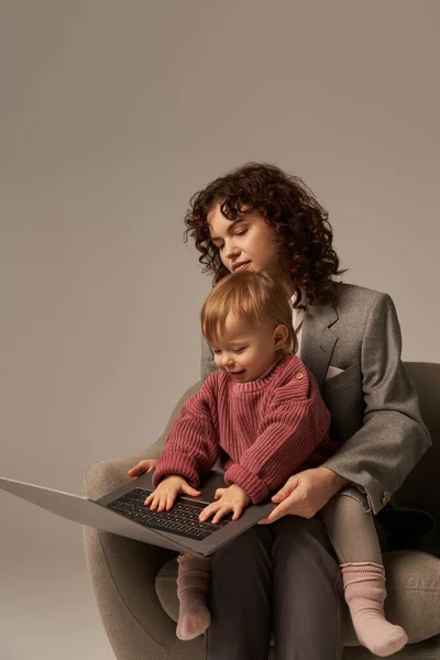 Построение успешной карьеры, баланс трудовой жизни, работающий родитель, кудрявая деловая женщина с помощью ноутбука и сидя на кресле с дочерью, матерью и ребенком, материнство, многозадачность — стоковое фото