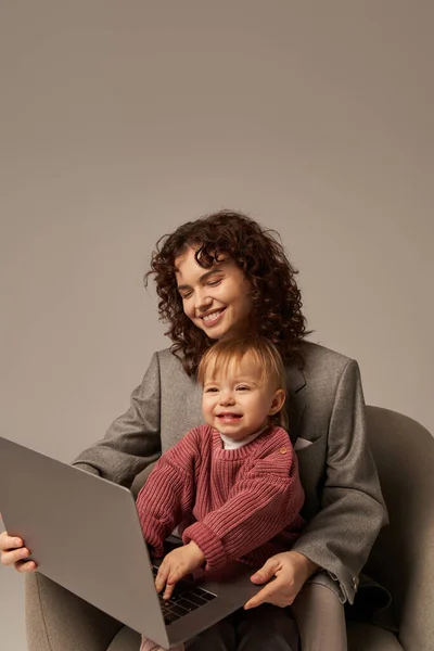 Construção de carreira bem sucedida, estilo de vida equilibrado, pai trabalhando, mulher encaracolada feliz usando laptop e sentado em poltrona com filha, mãe e filho, maternidade, multitarefa — Fotografia de Stock