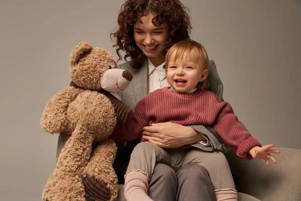 Quality family time, parenting and career, cheerful businesswoman holding teddy bear near toddler daughter on grey background, sitting on armchair, work life harmony concept — Stock Photo