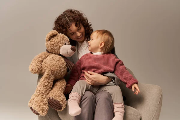 Quality family time, parenting and career, happy businesswoman holding teddy bear near toddler daughter on grey background, sitting on armchair, work life harmony concept, working parent — Stock Photo