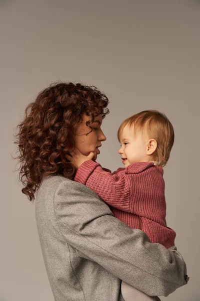 Working mother, parenting and career, curly businesswoman holding in arms her toddler daughter on grey background, work life harmony concept, loving motherhood, quality time, side view — Stock Photo