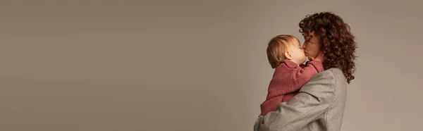 Elternschaft und Karriere, lockige Geschäftsfrau, die sich in den Armen hält und ihre kleine Tochter auf grauem Hintergrund küsst, Harmoniekonzept für das Arbeitsleben, liebevolle Mutterschaft, Qualitätszeit, Seitenansicht, Banner — Stockfoto
