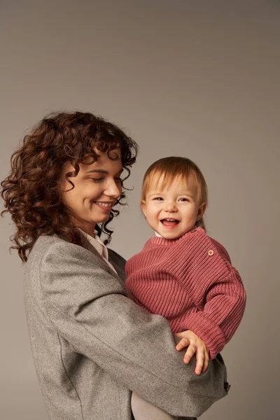 Quality family time, parenting and career, happy working mother holding in arms her toddler daughter on grey background, work life harmony concept, curly woman, loving motherhood — Stock Photo