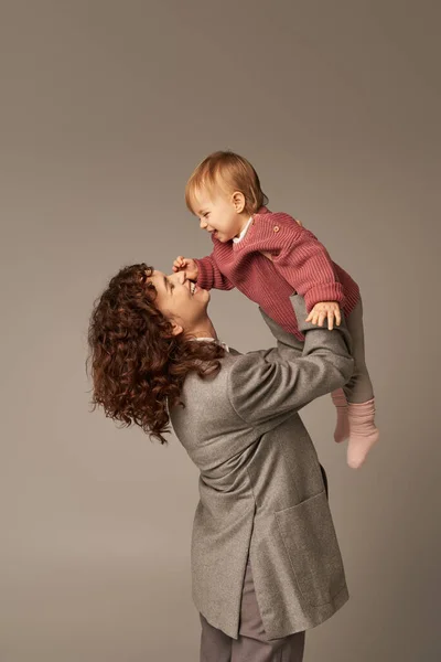 Работающая мать, воспитание и карьера, веселая деловая женщина, поднимающая свою дочку на сером фоне, концепция гармонии трудовой жизни, любящее материнство, качественное семейное время — стоковое фото