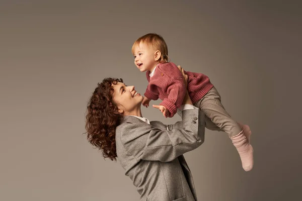 Positividade, mãe trabalhadora, parentalidade e carreira, empresária alegre levantando sua filha criança feliz em fundo cinza, conceito de harmonia na vida profissional, maternidade amorosa, tempo de família de qualidade — Fotografia de Stock