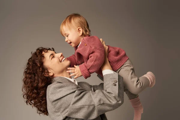 Madre trabajadora, crianza y carrera, mujer de negocios rizada levantando en brazos a su hija pequeña sobre fondo gris, concepto de armonía de la vida laboral, maternidad amorosa, tiempo familiar de calidad - foto de stock