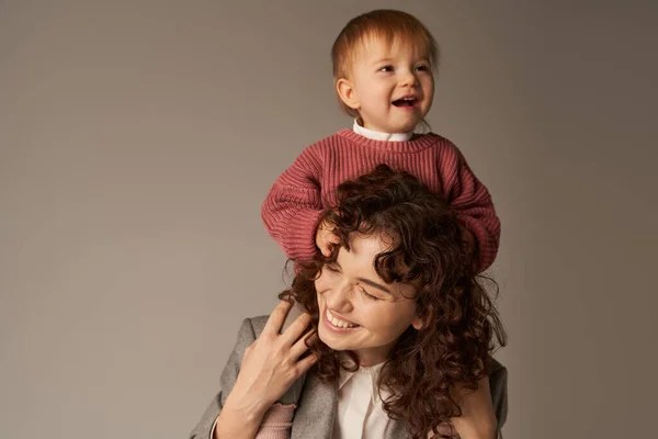 Motherhood, kid on shoulders, work life balance, curly businesswoman and happy daughter on grey background, work life harmony concept, loving motherhood, quality family time — Stock Photo