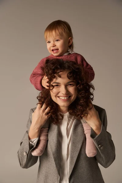 Loving motherhood, parenting and career, curly businesswoman and happy toddler daughter on grey background, kid on shoulders, work life harmony concept, loving motherhood, quality family time — Stock Photo