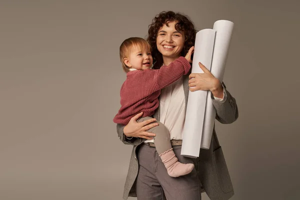 Mujer multitarea, logros profesionales, gestión del tiempo, madre alegre sosteniendo en brazos hija y rodado planos de papel sobre fondo gris, la construcción de una carrera exitosa - foto de stock