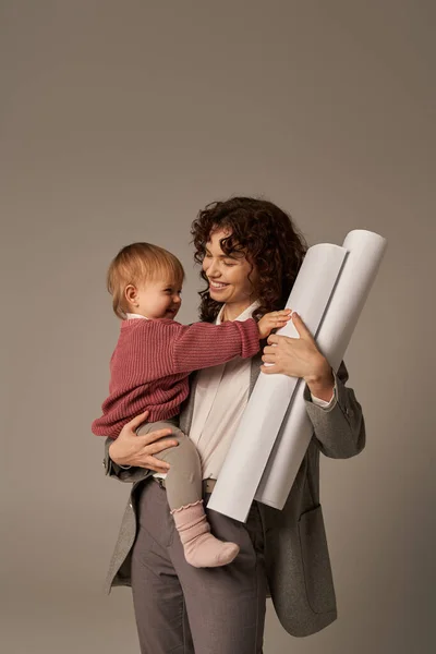 Aufbau einer erfolgreichen Karriere, Multitasking-Frau, berufliche Errungenschaften, Zeitmanagement, glückliche Mutter mit Tochter im Arm und gerollten Papierentwürfen auf grauem Hintergrund — Stockfoto