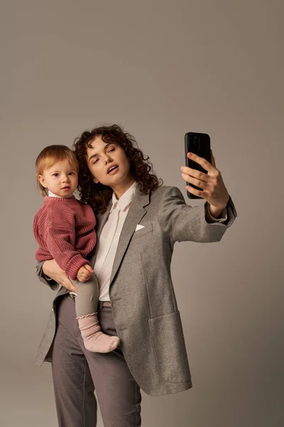 Modern parenting, building successful career, empowered woman, curly mother taking selfie on smartphone and holding in arms toddler daughter on grey background, motherhood — Stock Photo
