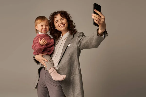 Modern parenting, work life balance, empowered woman, happy mother taking selfie on smartphone and holding in arms toddler daughter on grey background, motherhood — Stock Photo