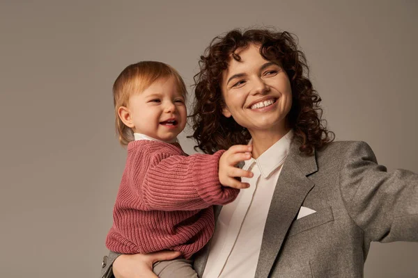 Career and family, motherhood, quality family time, happy and curly woman holding in arms toddler daughter on grey background, balancing between work and life, positivity — Stock Photo