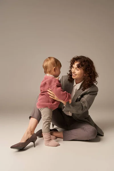 Career and family, balancing between work and life, cheerful woman with curly hair hugging toddler daughter on grey background, quality time, modern parenting, motherhood, full length — Stock Photo
