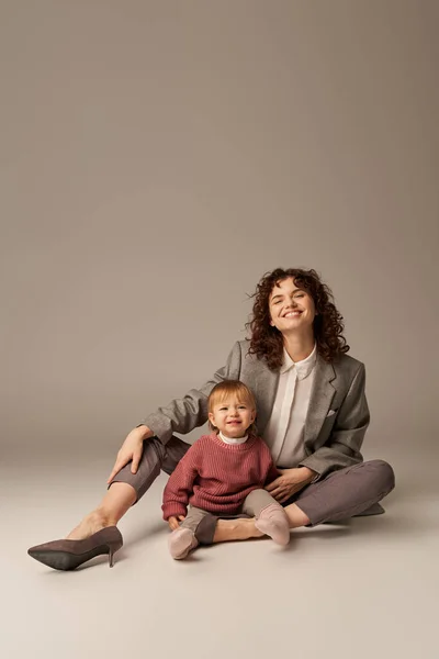 Career and family, balancing between work and life, happy woman with curly hair hugging toddler daughter on grey background, quality time, modern parenting, motherhood, full length — Stock Photo