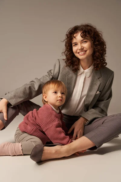 Career and family, balanced lifestyle, cheerful mother with curly hair hugging toddler daughter on grey background, quality time, modern parenting, businesswoman, loving motherhood — Stock Photo
