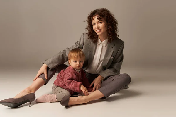 Parenting and family, balanced lifestyle, cheerful mother with curly hair hugging toddler daughter on grey background, quality time, modern woman, businesswoman, loving motherhood — Stock Photo