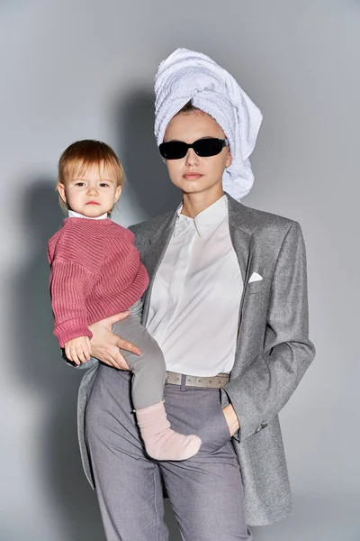 Multitasking, woman in sunglasses holding in arms toddler daughter and standing with towel on head, balancing between work and life, empowered woman in formal wear on grey background — Stock Photo