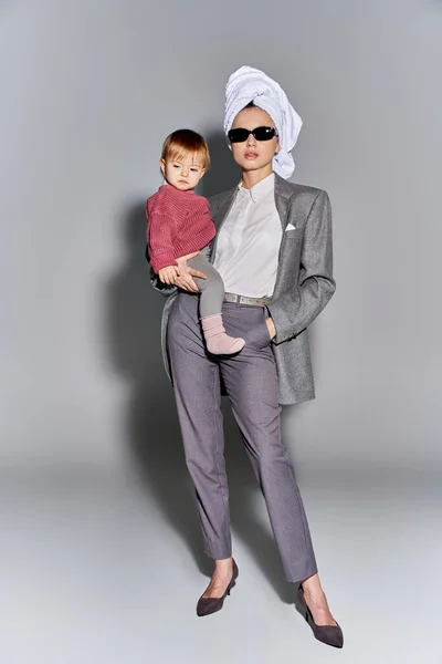 Багатозадачність, жінка в сонцезахисних окулярах тримає в руках дочку малюка і стоїть з рушником на голові, балансуючи між роботою і життям, потужна жінка в офіційному одязі на сірому фоні, повна довжина — стокове фото