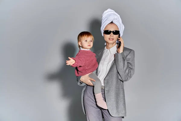 Multitasking, woman in sunglasses holding in arms toddler daughter and standing with towel on head, balancing lifestyle, businesswoman in formal wear talking on smartphone on grey background — Stock Photo
