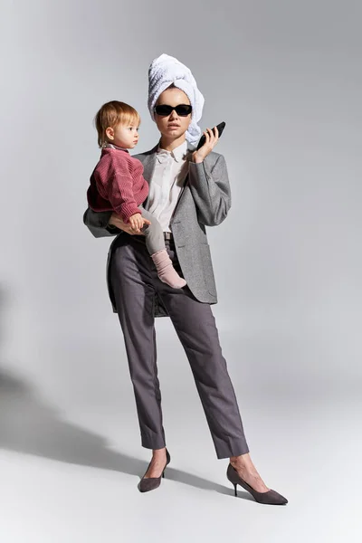 Multitasking, woman in sunglasses holding in arms daughter and standing with towel on head, balancing lifestyle, businesswoman in formal wear holding smartphone on grey background, full length — Stock Photo