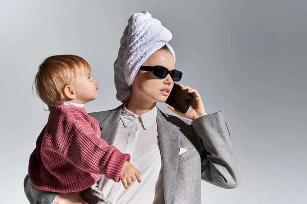 Zeitmanagement, Multitasking, Frau mit Sonnenbrille, Tochter im Arm und mit Handtuch auf dem Kopf, Geschäftsfrau in offizieller Kleidung, die auf Smartphone vor grauem Hintergrund spricht — Stockfoto