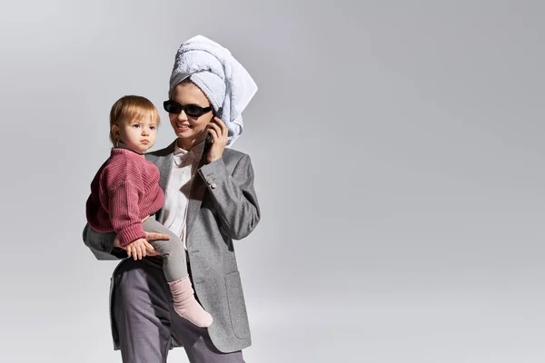 Wachstum und Familie, Frau mit Sonnenbrille, Tochter im Arm und mit Handtuch auf dem Kopf, balancierender Lebensstil, Geschäftsfrau in offizieller Kleidung, die auf Smartphone auf grauem Hintergrund spricht — Stockfoto