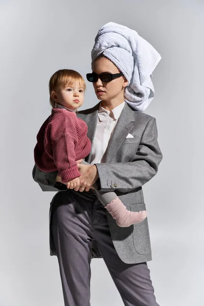 Mulher em óculos de sol segurando em braços criança filha e de pé com toalha na cabeça, multitarefa, equilíbrio entre trabalho e vida, mulher empoderada em desgaste formal em fundo cinza — Fotografia de Stock