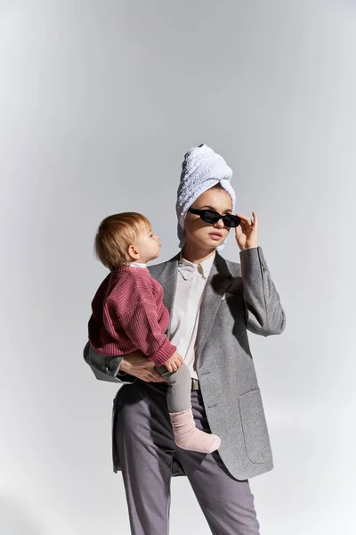 Woman in sunglasses holding in arms toddler daughter and standing with towel on head, multitasking, balancing lifestyle, empowered woman in formal wear on grey background — Stock Photo