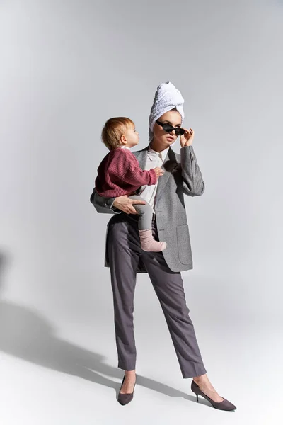 Femme dans les lunettes de soleil tenant dans les bras tout-petit fille et debout avec serviette sur la tête, multitâche, mode de vie équilibré, femme habilitée en tenue formelle sur fond gris, pleine longueur — Photo de stock