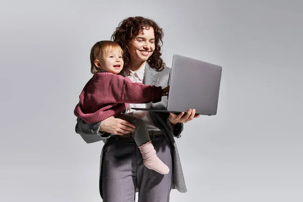 Time management, working mother holding laptop and toddler daughter in arms on grey background, work life harmony, career and family, modern parenting, professional success, cheerful businesswoman — Stock Photo