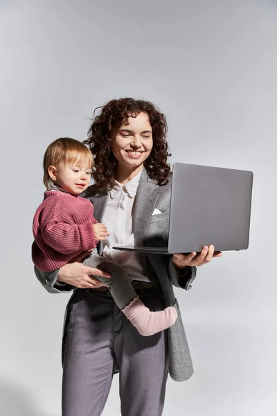 Gestión del tiempo, madre trabajadora feliz sosteniendo el ordenador portátil y la hija del niño en brazos sobre fondo gris, armonía de la vida laboral, carrera y familia, crianza moderna, éxito profesional, mujer de negocios - foto de stock