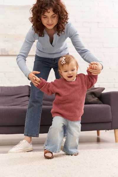 Перші кроки, якісний час, зв'язок, балансування роботи і життя, сімейні відносини, робітнича мати тримає руки з дочкою малюка, спокій, затишна вітальня, джинсові джинси, повсякденний одяг — стокове фото