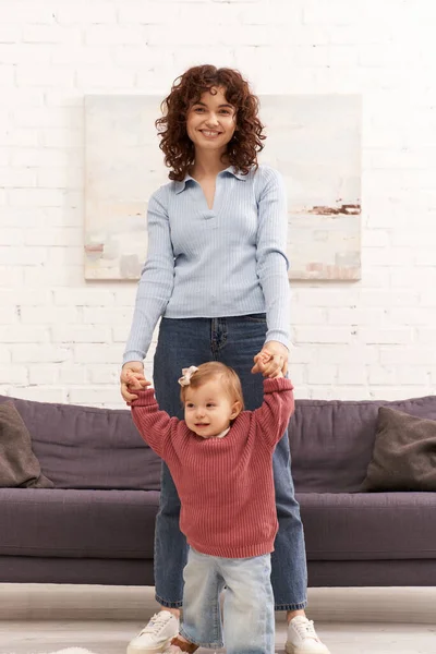 Перші кроки, якісний сімейний час, зв'язок, балансування роботи і життя, щаслива робоча мати тримає руки з дочкою малюка, спокій, затишна вітальня, джинсові джинси, повсякденне вбрання — стокове фото