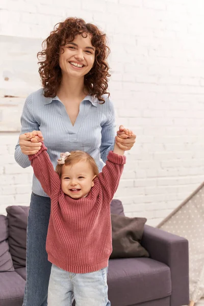 Loving motherhood, balancing work and life, family bonding, happy and curly mother lifting toddler daughter, togetherness, cozy living room, quality time, denim jeans, engaging with child — Stock Photo