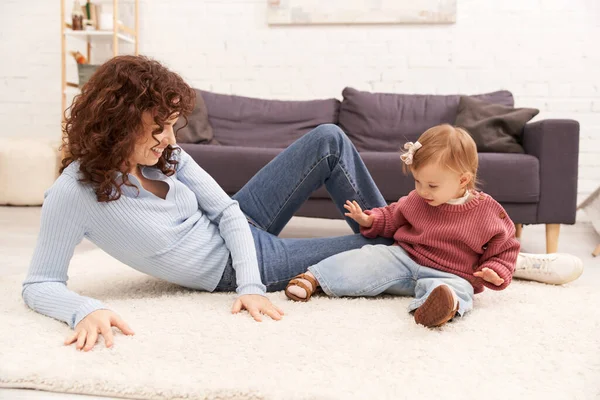 Work life balance, happy and curly woman sitting on carpet with excited toddler daughter in cozy living room, quality family time, casual attire, bonding between mother and child — Stock Photo
